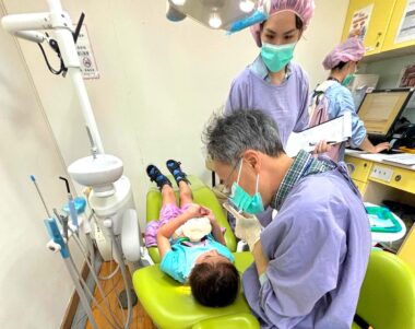 A 6-year-old girl lies prone in a dentist's chair as an older male dentist leans over her explaining things to her. Two dental assistants are working nearby. The dentist's office looks like a typical dentist's office, with lime green chairs and cabinets. The photo is taken from behind the dental chair and dentist, down at the top of the girl's head, which is a dynamic angle for the photo and projects awkwardness and activity.