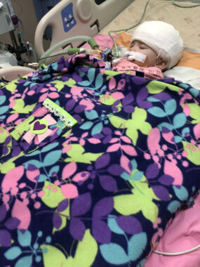 A baby whose head is wrapped in a large gauze bandage rests in a hospital bed, atop a pink sheet and under a multicolored blanket, which has a field of black almost completely covered by blue, purple, pink, and green designs.