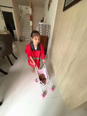 A mesmerizing photo with weird angles and distorted object ratios from the inside of a home. In the center of the photo is a young girl pushing toy poodle puppy around in a pink doll stroller.