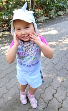 A young girl, maybe 5 or 6 years old, smiles for a photo with her hands on her cheeks. She's wearing a T-shirt, shorts, sparkly sandals, and socks, as well as a hat with a unicorn horn poking out the top. There's a colorful handkerchief tied around her neck to accommodate her excessive drooling. She's standing on a shaded stone path outside, with dense foliage visible behind her.