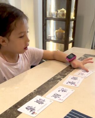 A young girl sits at an Asian-style dinner table and points to one of a series of cards on the table. The cards have Chinese symbols on them and are used in learning Chinese.