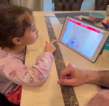 A young girl points and looks at an iPad sitting on the dinner table. 