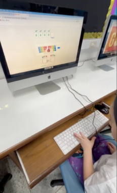 We see a white desktop, on which sits a desktop computer. Its keyboard is below on a slide-out tray. A child's fingers are on the keyboard. To the right we can just see that there's another desktop computer.