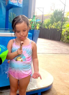 A 6-year-old girl eats a treat while wearing a blue and pink swimsuit. She's standing outside in front of a small blue building, perhaps where the treats are sold, and there is lush vegetation visible beyond the path behind her.