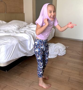 A young girl, maybe 5 or 6 years old, giggles while pulling a purple shirt over her head. She has on blue patterned leggings and is standing on a wooden floor next to a bed.