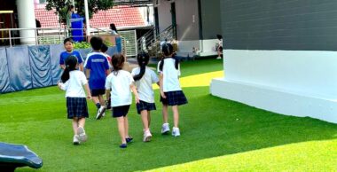 A group of four young girls – first graders – are seen walking away, their backs toward the camera. They're walking on what appears to be artificial grass outside of their school building, and several boys stand farther away. The girls are all in matching uniforms of white shirts and dark skirts.