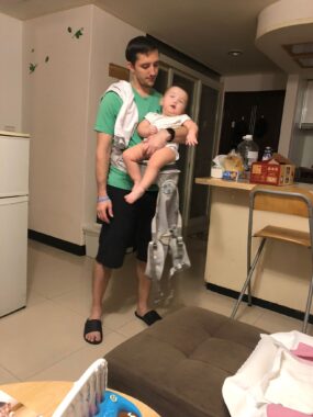 A man holds a baby girl in his left arm while standing in a small apartment looking down at the floor. He's in dark shorts and a green T-shirt, with a baby towel draped over his shoulder. 