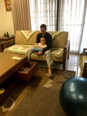 A man sits on a couch in a living room, with a baby sitting upright in his lap. The room is in shades of brown, and a window with vertical shades is behind them.