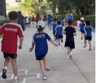 A group of children run down a paved walkway as part of a school walk-a-thon. Most are wearing blue or red shirts with the school's name on the back. The path is lined by green hedges and there are palm trees in the distance.