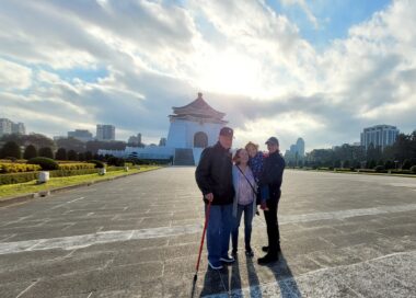 An outdoor photo of a couple, their baby, and another man with a cane. Dramatic, long angles highlight a sunny sky with dynamic cloud formations, as well as the long shadows. 