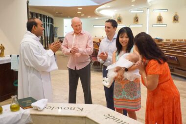 Four people and a newborn attend a baptism ceremony with the fifth person, who is administering it. They are in the sanctuary of a church.