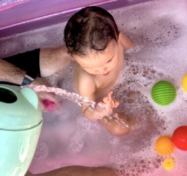 An overhead shot shows a baby girl sitting in a bathtub filled with bubbles. We can see her dad's leg behind her, and he appears to be pouring water out of some sort of bucket. The girl is holding up her right hand to touch the stream of water. There are several colorful balls floating on the surface of the water in the tub, and the bath appears to be lit up pink.