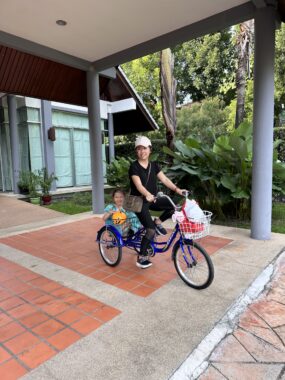 A woman and her daughter pause for a photo on a covered walkway outside their home. The mom is pedaling a blue tricycle while the girl, who's about 6 years old and wearing an Elsa dress, sits in a large basket in the back. They're preparing to go trick-or-treating, as part of their adapted Halloween celebration.