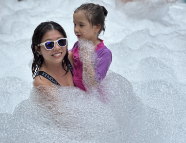 A woman smiles while holding up her 6-year-old daughter at a foam party. They both look to be wearing bathing suits, and the woman has on a pair of white-framed sunglasses. They are completely surrounded by foam, which comes up nearly to the woman's shoulders.