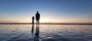 A low-angle photo shows the silhouette of a man and his young daughter standing on the beach holding hands. The sun is setting in the distance.