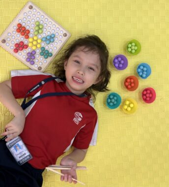 An overhead shot of a young girl smiling and lying on her back on a yellow children's mat. Around her head are colorful educational children's games. 