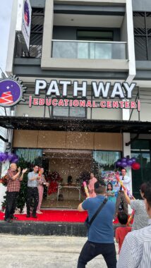 A crowd of people stands outside a building with a sign that reads "Pathway Educational Center." There's a red carpet outside the front doors and some clusters of balloons to celebrate the center's opening. Everyone is clapping and cheering.