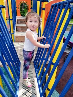 A toddler girl stands on a narrow walkway that's part of a playground structure. There are vertical blue bars on either side of the walkway, which she's holding on to. Her body is facing one side of the bars, as if looking out over the playground, but her head is turned to her right side as she looks at the camera.