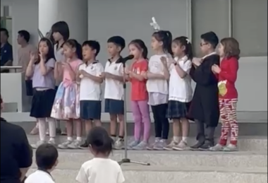 A line of 10 children, with a microphone stand around the middle, stand on what appear to be concrete steps. Two or three audience members appear in the bottom left foreground.