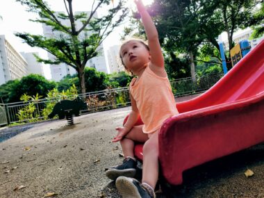 A young girl in a peach tank top sits on a red plastic slide and reaches upward. She appears to be in a playground, with trees in the background.