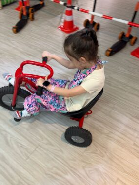 A young girl, about 6 years old, pedals a red tricycle on an indoor bike track. The shot is taken from above, from the perspective of an adult standing next to her, and the girl is looking down in concentration.
