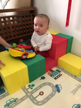 A toddler, with little hair and wearing a white shirt, sits on a red soft block, with a couple in front of her and a couple behind her. A toy is before her fingertips, and the rug features pictures of a road, cars, and buildings.