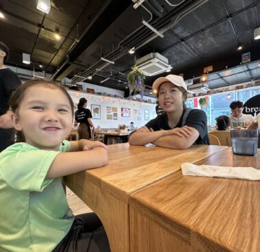 A woman and her daughter, who's about 6 years old, sit across from each other at a table in a restaurant. The photo is taken from a low angle, about the height of the table, and several other tables and people are visible in the background. Both the woman and her daughter have their arms crossed on the table in front of them and are grinning at the camera.