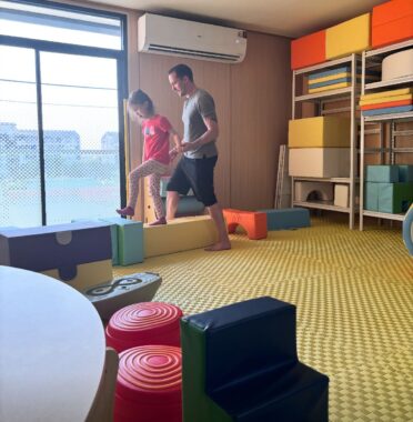 A young child in a red T-shirt and tan pants stands on a yellow soft block, about to step forward onto a smaller green block. A barefoot man with short dark hair, a gray T-shirt, and blue shorts stands behind her, with a hand near her hip. The room what appears to be a rubber yellow floor, and there are other such blocks nearby as well as on some shelves.