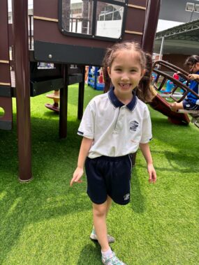 A young girl with light brown hair in pigtails faces the camera and smiles as she stands on what appears to be green turf. She wears a white short-sleeve shirt with a blue collar and insignia and blue shorts, something like a school uniform. Behind her is what appears to be a brown playhouse on short stilts.