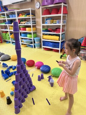 A young girl in a pale violet tank top and pink skirt looks at a tower of purple cups, broad at the bottom and narrow at the top. She's on a yellow floor with playthings and other cups nearby, as well as shelves of other colorful items.