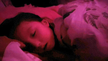 A young girl is fast asleep in bed, covered by sheets. A pink hue from the light glows throughout the photo.