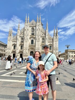 A young girl poses for a photo with her dad and her grandmother in front of the Duomo cathedral in Milan, Italy. The girl is wearing a pink and orange dress and a baseball cap, and she looks to be holding a water bottle. It looks like a sunny day, with only a few wispy clouds in the blue sky.