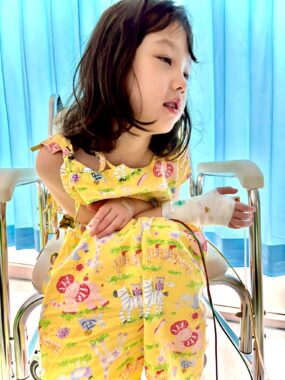 A young girl, with a bandaged hand and a couple of visible tubes, sits on what appears to be a portable seat in front of a bluish-green curtain.