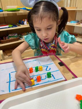 A young girl in pigtails sits at a table and works with manipulatives and a whiteboard that has numbers in boxes. Shelves with brightly covered objects are behind her.