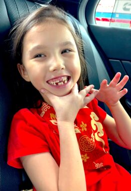 A young, smiling girl grasps her chin and shows off a mouth of missing teeth, baby teeth, and a couple adult teeth. She's sitting in the back seat of a car.