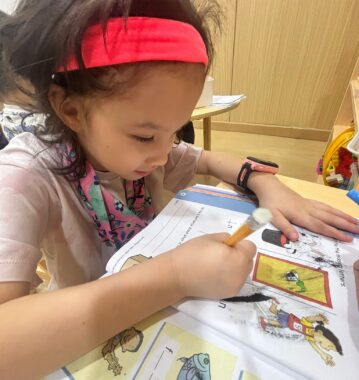 A young girl wearing a red headband, a white T-shirt, and a multicolored bandana around her neck is writing in a workbook.