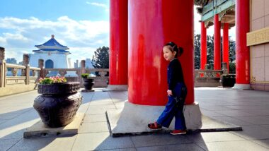 A young girl stands by a very large round column outside a building. In the distance is a fence and, beyond that, what appears to be an Asian temple.