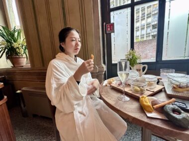 A woman wearing a robe sits at a table by a window, enjoying a meal.