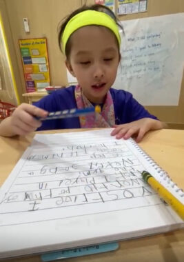 A young girl, maybe 6 or 7 years old, sits at a table working on a writing assignment. She's holding a pencil in her right hand and looking at a notebook in front of her.