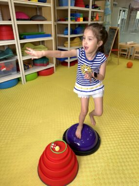 A young girl in white shorts, a blue-and-white-striped shirt, and bare feet stands on one foot on a purple balance ball. The floor is a yellow mat; white shelves along a wall nearby contain similar brightly covered objects and other items.