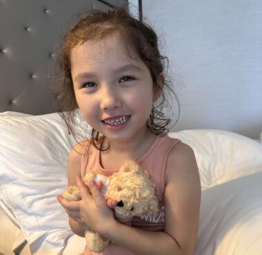 A young girl sits smiling on a bed, clutching a small teddy bear.