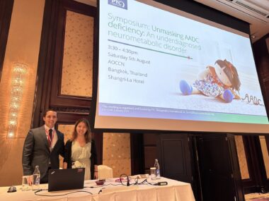 Two people stand behind a table under a large screen at a conference.