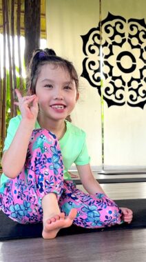 A young girl, about 6 or 7 years old, sits on a black yoga mat in a yoga studio. She's smiling at the camera and making a peace sign with her right hand. 