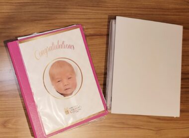 Two binders are sitting on a wooden desk or table. The one on the right is plain white, and the one on the left is pink, with a photo of a baby's face in the center with the word "Congratulations" above it.