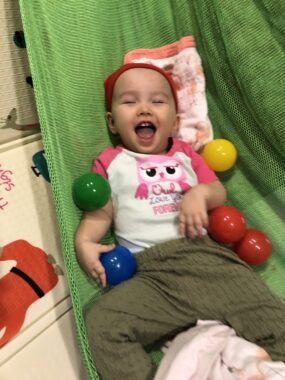 A young child lying in a hammock laughs as she plays with a selection of brightly colored, plastic balls.
