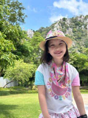 A young girl smiles happily while standing outdoors on a sunny day, surrounded by mountainous and green landscapes. She's wearing brightly colored, pink-themed clothing, including a very stylish fedora. 