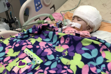 A small child is shown in a hospital bed, under a blanket, preparing for AADC deficiency gene therapy.