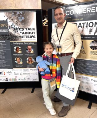 A man poses for a photo with his daughter in front of a few large posters at a conference. He's wearing a lanyard and holding a tote bag that says "AADC Trust," and his daughter, who looks to be about 7 years old, is wearing a rainbow striped sweater and holding a stuffed toy.