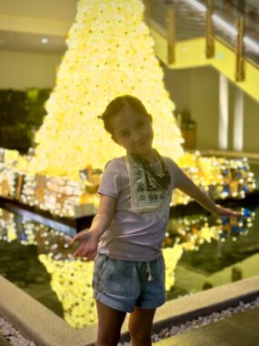 A young girl stands in front of a Christmas tree and decorations.
