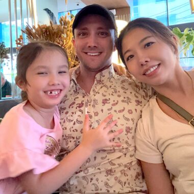 A family of three — husband, wife, and young daughter — lean in close and smile for a photo.